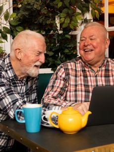 Two men smiling and laughing as they drink coffee in front of a laptop